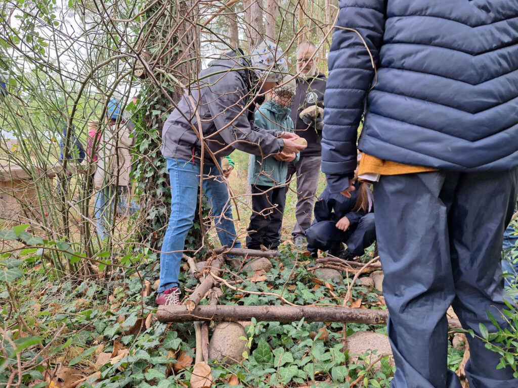 Der Umriss der Futterhütte wurde mit Steinen und Ästen markiert. Doris erklärt welche Steine als nächstes gebraucht werden.