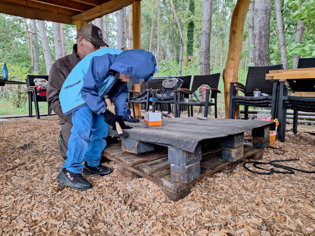 Ein Junge hämmert die Dachpappe für das Futterhaus an einer Europalette fest. Im Hintergrund passt ein Erwachsener auf, dass nichts passiert
